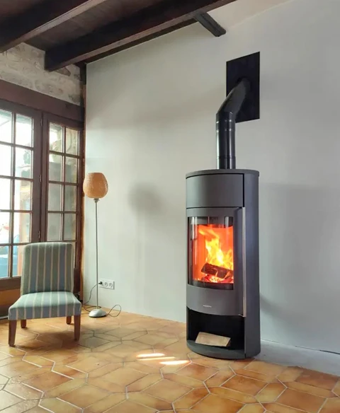 Poêle à bois design cylindrique noir mat installé dans un salon rustique aux poutres apparentes et carrelage tomette hexagonale