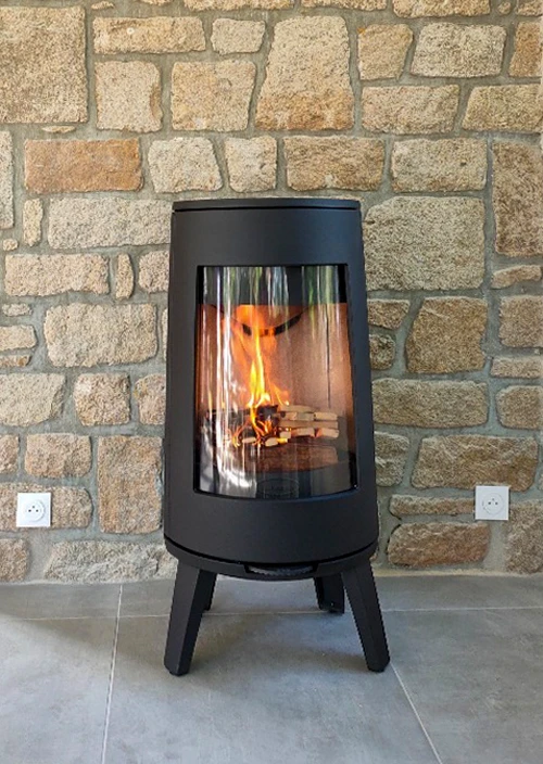 Poêle à bois design cylindrique noir mat, posé sur pieds métalliques tripodes, vitrage panoramique incurvé révélant un feu actif avec bûches en combustion